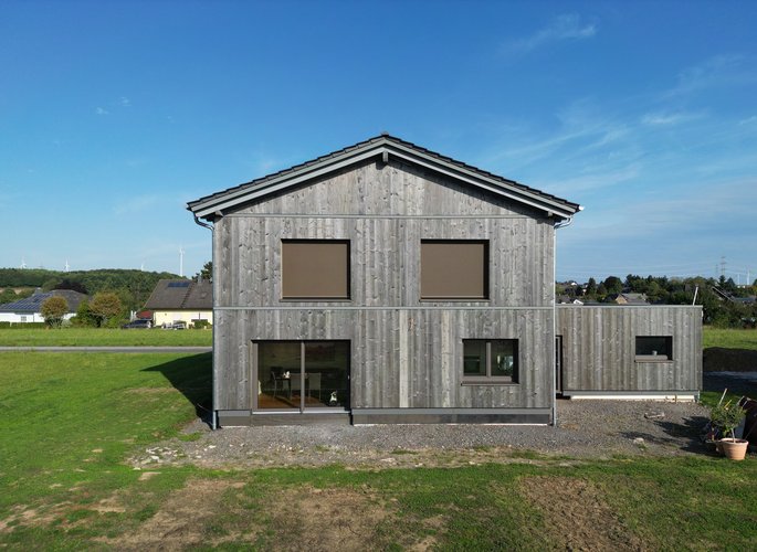 Frontalansicht eines modernen Holzhauses mit Flachdachanbau, geschlossenen Rollläden und umgeben von grüner Wiese unter blauem Himmel.