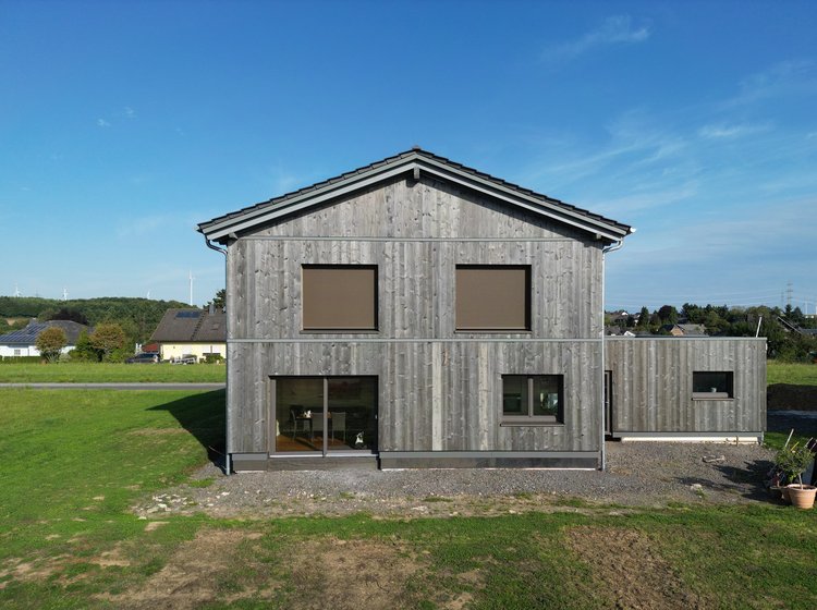 Frontalansicht eines modernen Holzhauses mit Flachdachanbau, geschlossenen Rollläden und umgeben von grüner Wiese unter blauem Himmel.