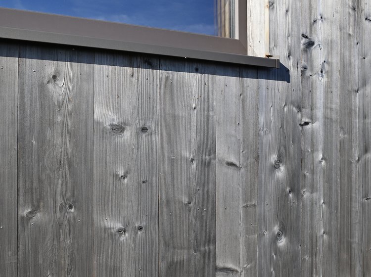 Detailansicht einer strukturierten, vertikalen Holzfassade mit Fenster und sichtbarer Maserung im Sonnenlicht.