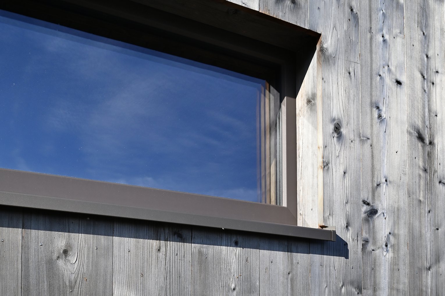 Detailansicht einer strukturierten, vertikalen Holzfassade mit Fenster und sichtbarer Maserung im Sonnenlicht.