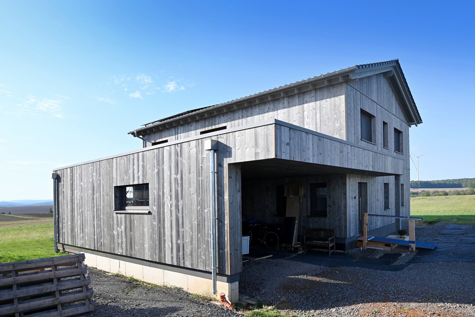 Zweistöckiges Holzhaus mit überdachtem Eingangsbereich, Carport und Kiesfläche, gelegen in ländlicher Umgebung bei klarem Wetter.