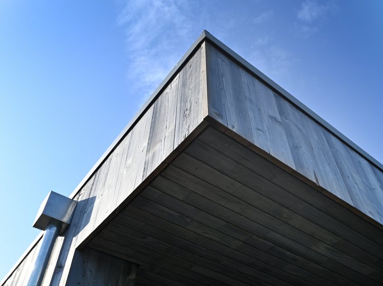 Nahaufnahme eines modernen Flachdachgebäudes mit vertikaler Holzverschalung und Metallregenrinne vor blauem Himmel.